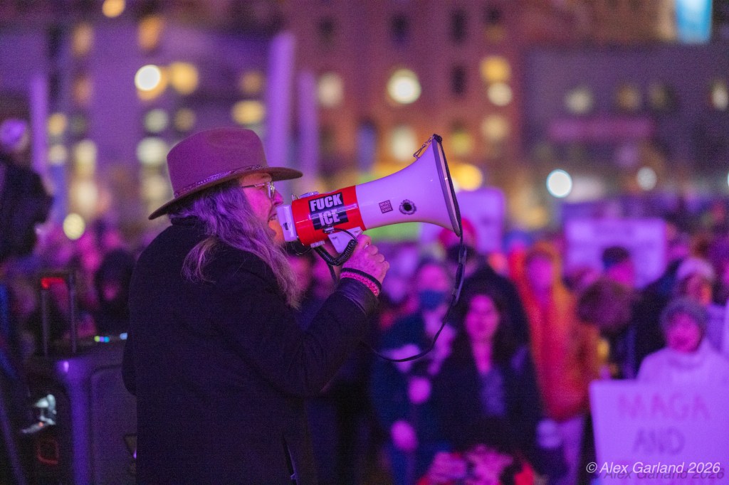 A person with long hair and a hat holds a megaphone with a provocative message while speaking to a crowd at a nighttime protest, illuminated by colorful lights.