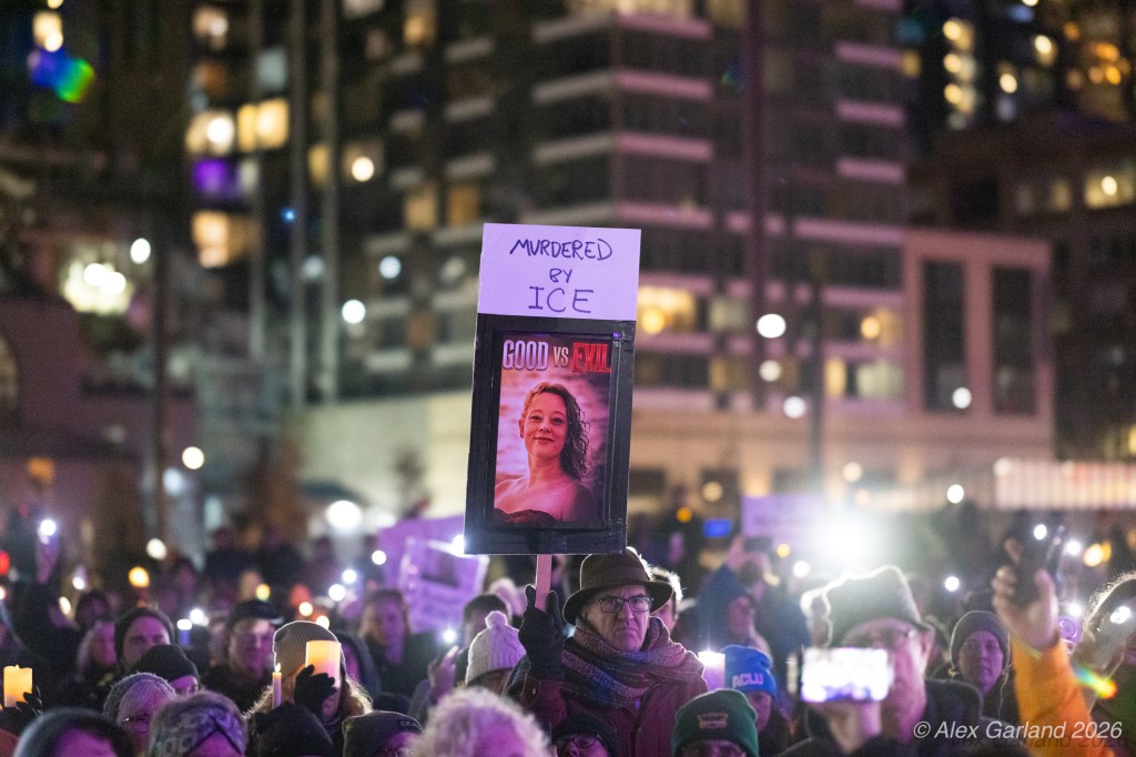 A crowd of protesters holding candles during a nighttime demonstration, with a person in the foreground holding a sign that reads 'Murdered by ICE' alongside an image of a woman.