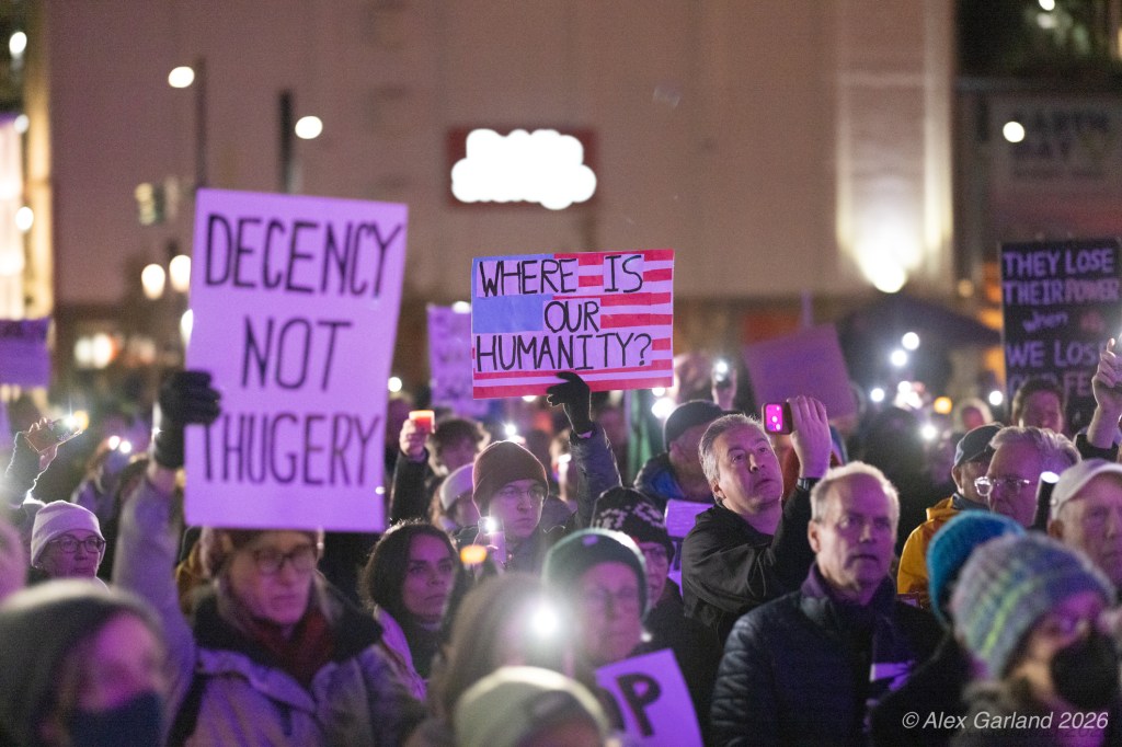 A large crowd of protesters holding signs in the evening, with some using flashlights or phone lights. Signs include messages like 'Decency Not Hugery' and 'Where is Our Humanity?'