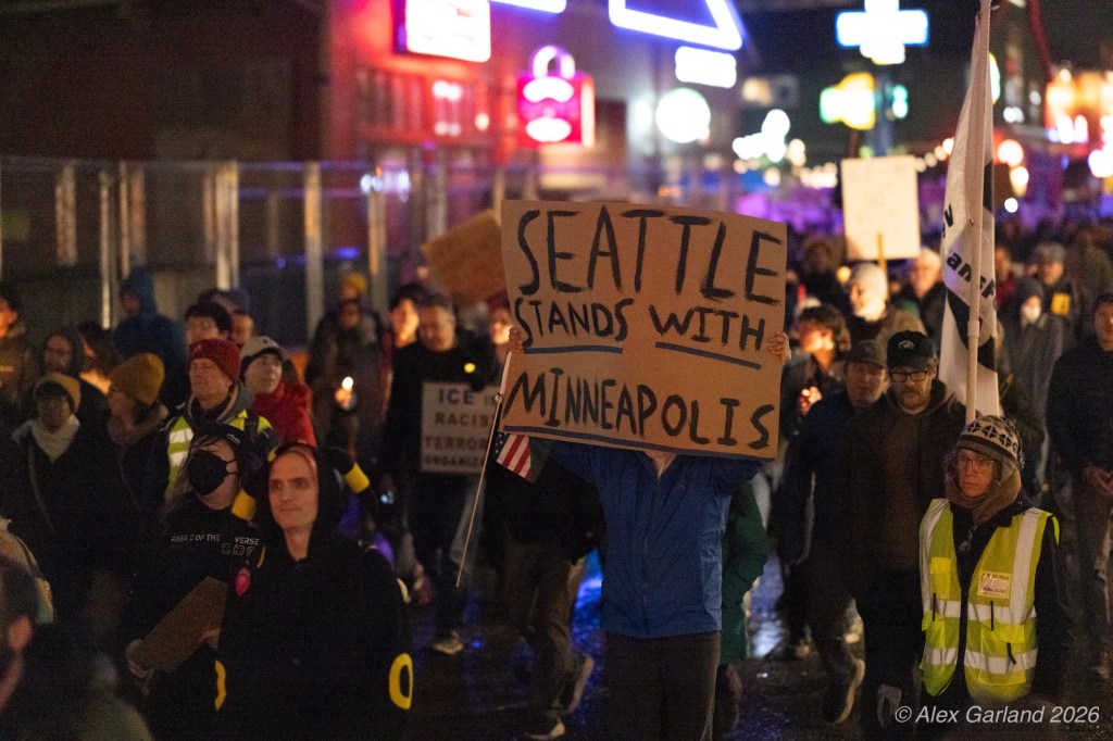 A nighttime protest with people holding signs, including one that says 'SEATTLE STANDS WITH MINNEAPOLIS.' Demonstrators are gathered in a city street with bright lights in the background.