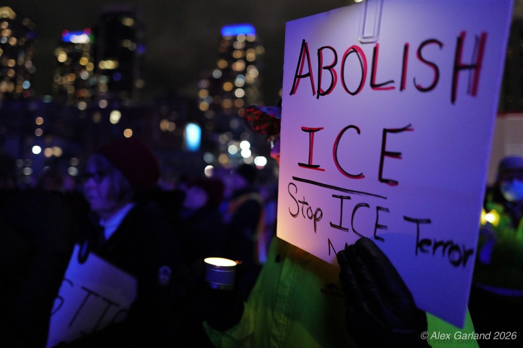 A protestor holding a sign that reads 'ABOLISH ICE' and 'Stop ICE Terror' during a nighttime rally with city lights in the background.