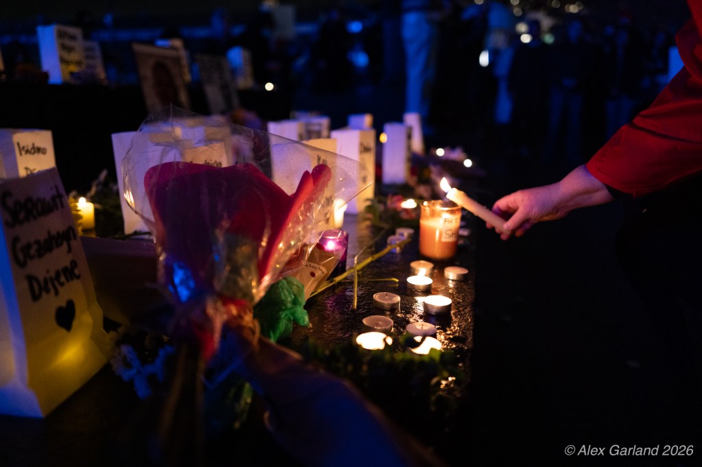 A person lights a candle at a memorial display featuring flowers and multiple candles, with handwritten signs in the background.