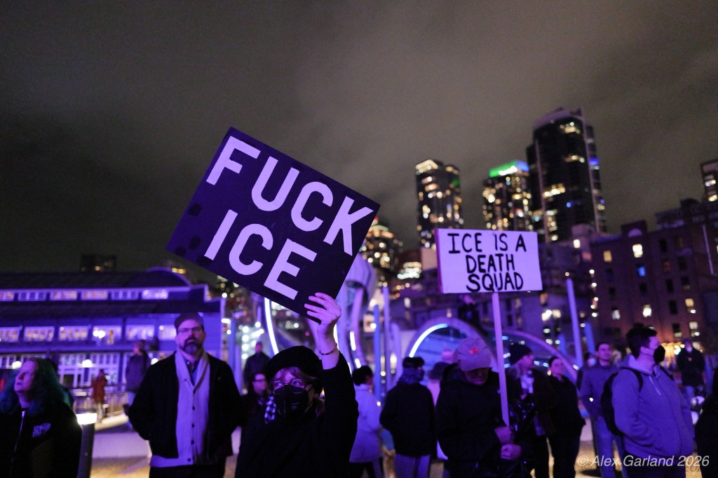 Demonstrators holding protest signs at night, with city skyline in the background.
