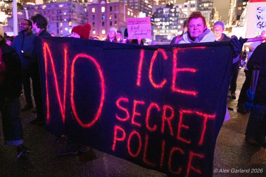 A crowd holding a large black banner with red text reading 'NO ICE SECRET POLICE' during a nighttime protest.