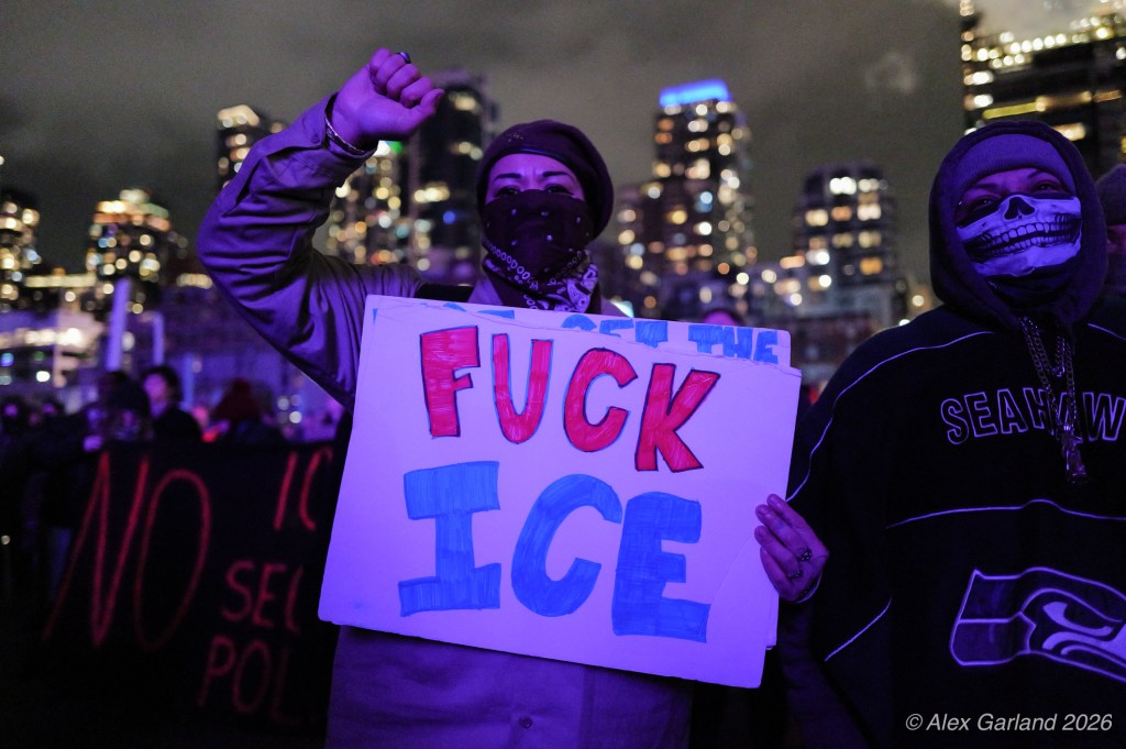 Two individuals at a night protest, one holding a colorful sign that reads 'F*** ICE', with city skyline in the background.