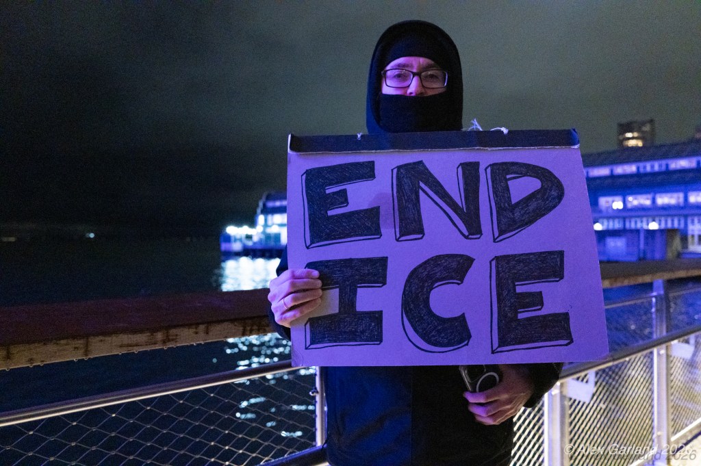 A person wearing a black face mask and glasses holds a large sign that reads 'END ICE' while standing by a body of water at night.