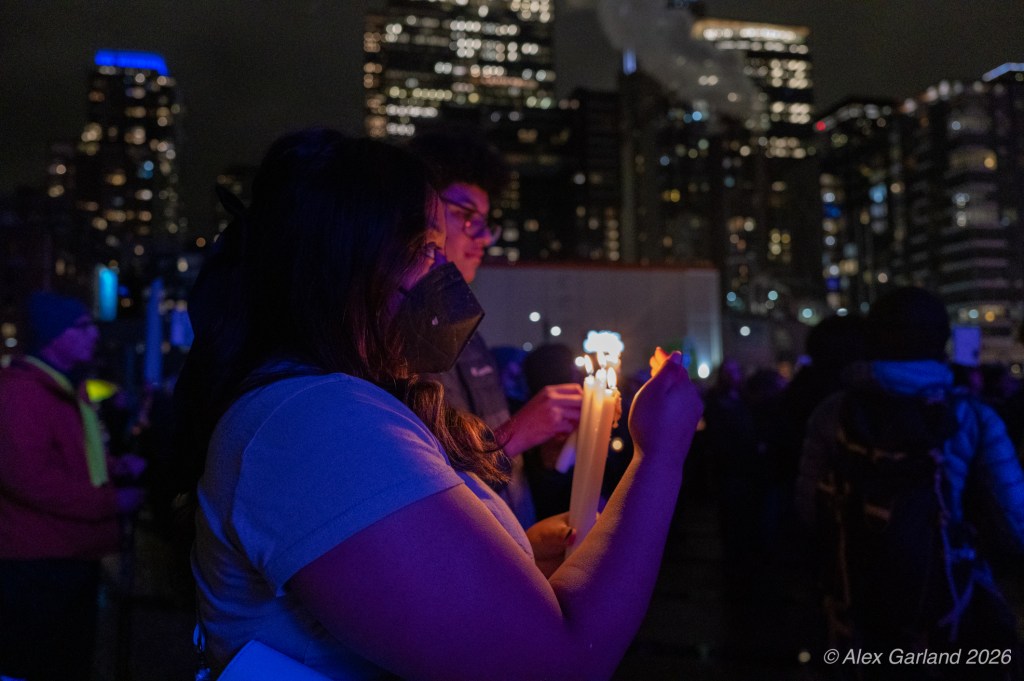 A woman holding lit candles in a dark urban setting, surrounded by others during a nighttime gathering with city lights in the background.