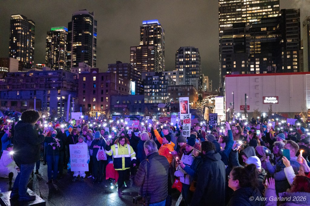 A large crowd gathered at night for a protest in an urban setting, with people holding signs and using mobile phone flashlights. A speaker is on a platform addressing the crowd, surrounded by high-rise buildings and city lights.