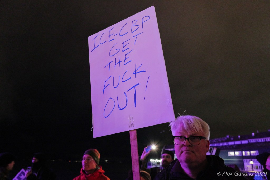 A protester holding a sign that says 'ICECBP GET THE FUCK OUT!' during a nighttime demonstration.