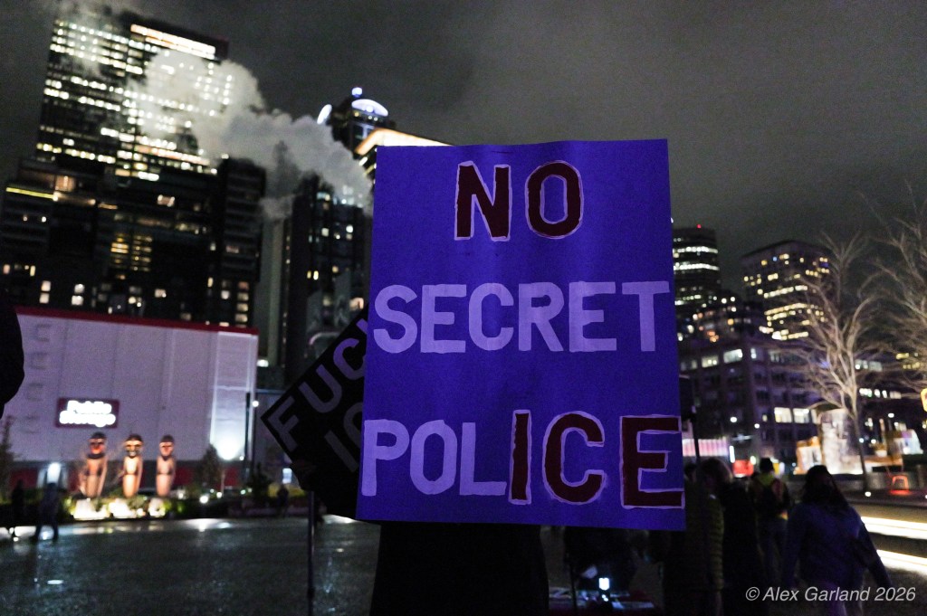 A protester holding a sign reading 'NO SECRET POLICE' in a city setting at night, with buildings and smoke in the background.
