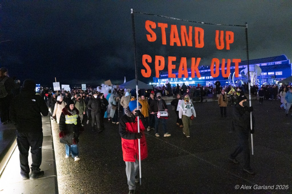 A crowd of protesters holding signs and a banner reading 'STAND UP SPEAK OUT' during a nighttime demonstration.