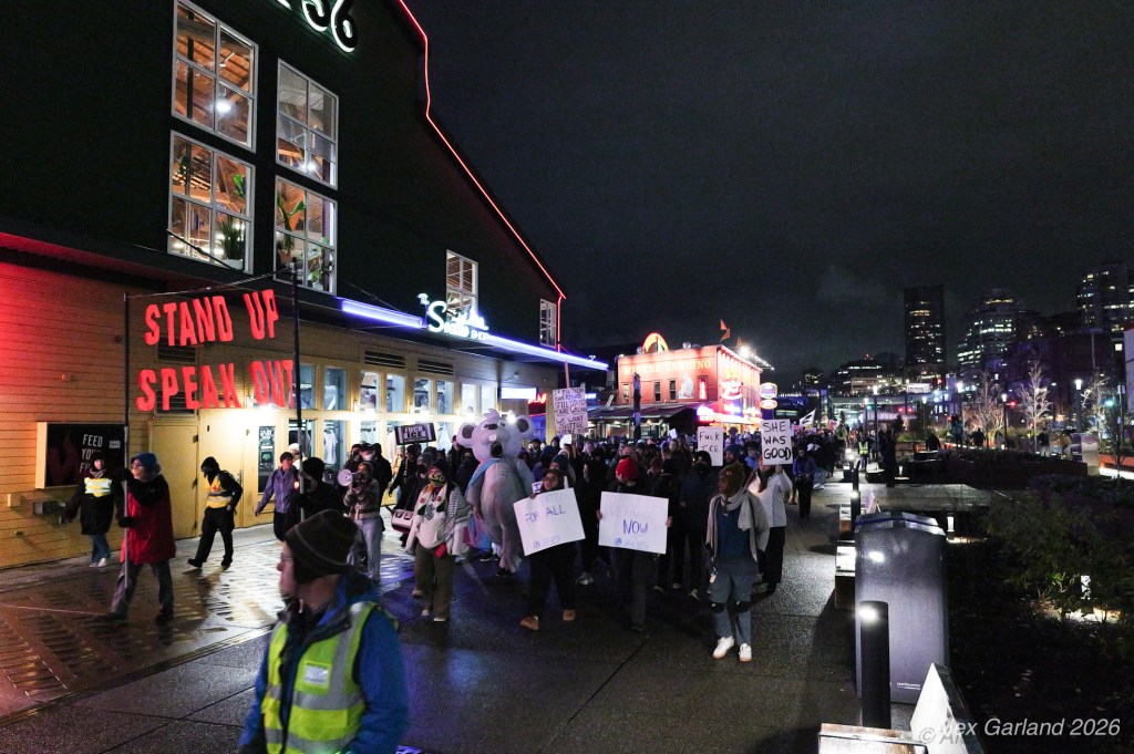 A nighttime protest scene in a city with people holding signs, including messages like 'Stand Up Speak Out' and 'She Was Good'. A large crowd is walking past a building illuminated with colorful lights.